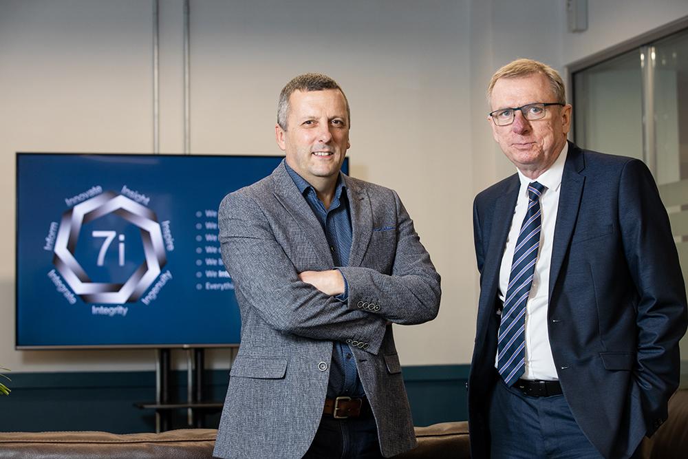 The founders of 7i Group standing in a meeting room with the company logo on the screen in the background