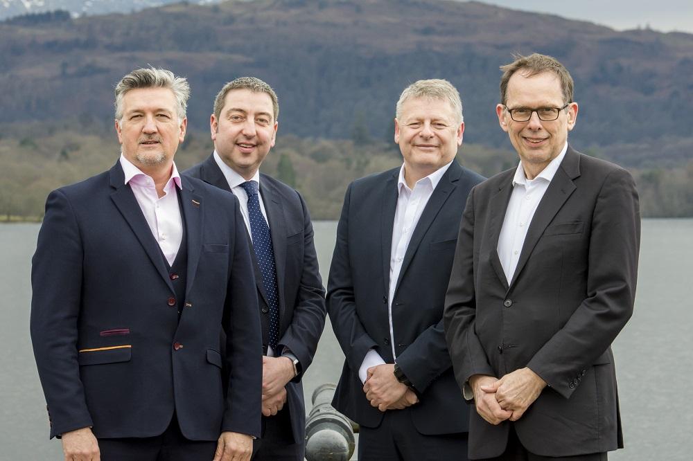 4 men in suits stood in front of a lake and hills