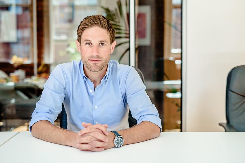 A man sat at a desk with a glass wall and office in the background