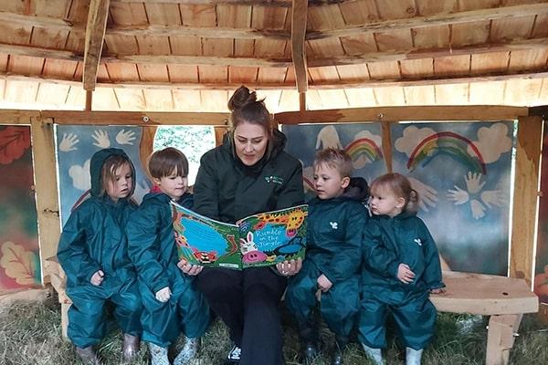 A member of nursery staff reading a book to four children, under the roof of a sheltered outdoor play area