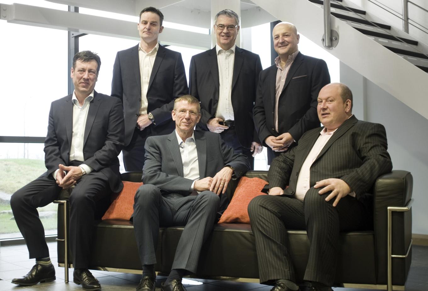 A group of men in suits sat and stood around a couch in an office building