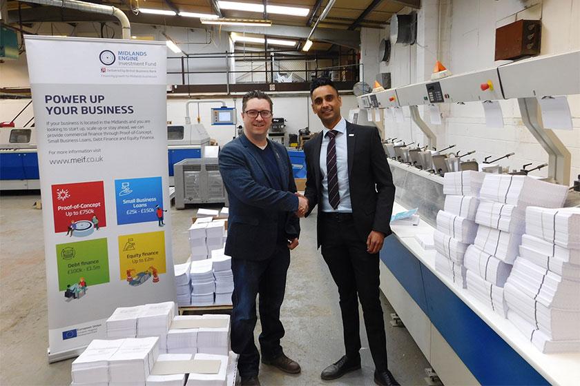 2 men shaking hands in a print lamination warehouse with stacks of paper besides them