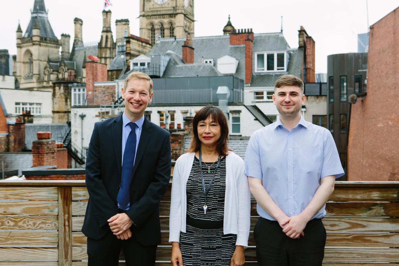 2 men and a woman stood on a balcony with buildings in the background