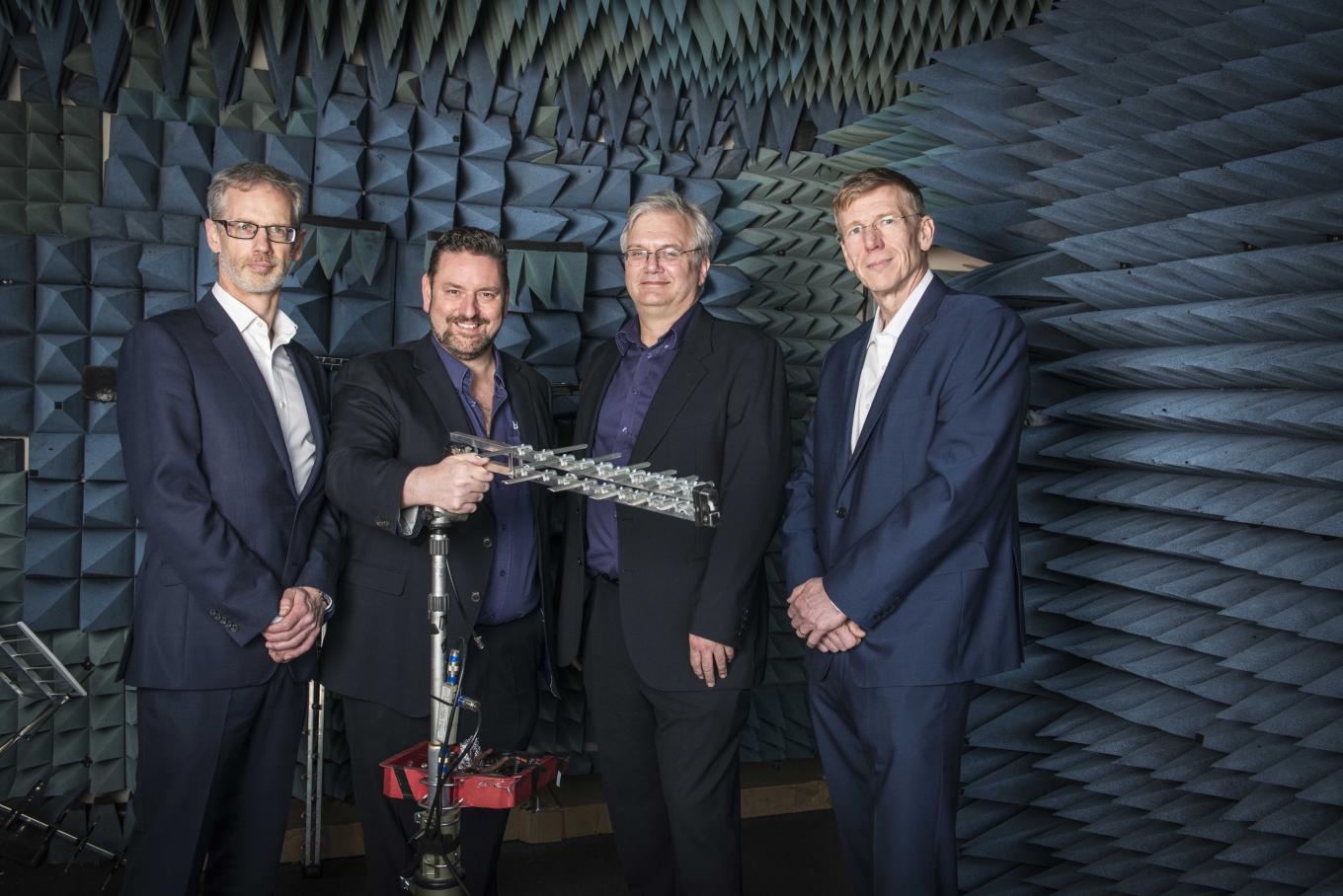 4 men in suits holding aerial equipment in a room with spikes on the wall