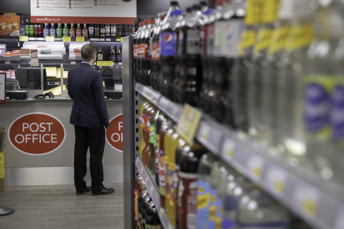 Shelves of fizzy drinks in a shop with a man at the post office counter