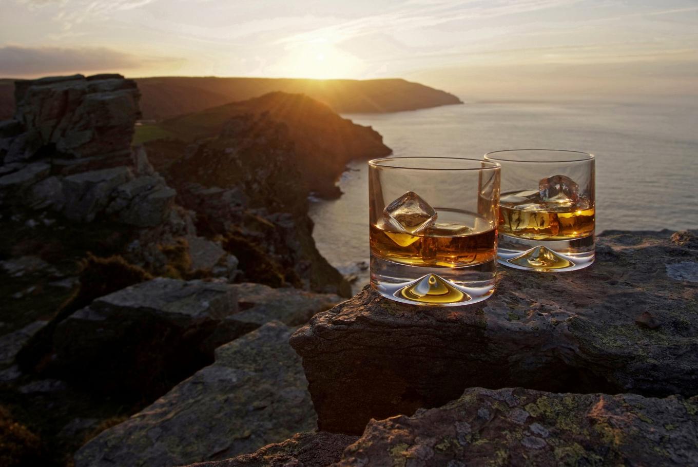 A glasses of whiskey with ice resting on a ledge overlooking the ocean and sunset
