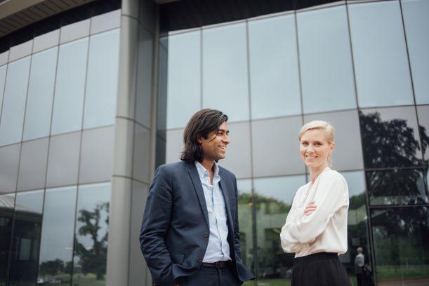 A man and a woman stood outside of a glass office building smiling