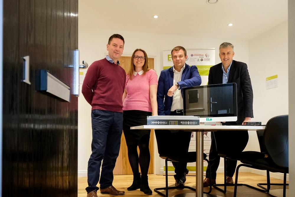 3 men and a woman stood in a meeting room around a table with a computer screen and books