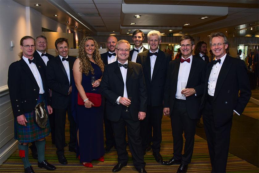 A group of men dressed in formal suits and bow ties and one woman in a maxi dress standing in a reception