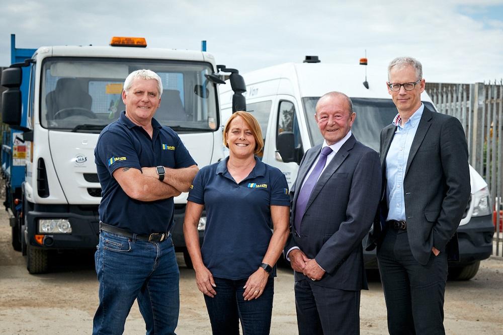 3 men and a woman smiling and stood outside in front of some vans