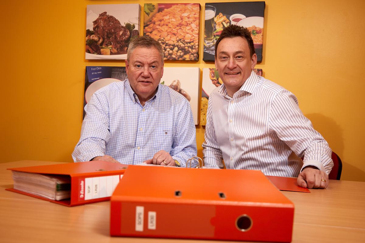 2 men sat at a table with 2 folders in front of them and images of food on the wall in the background