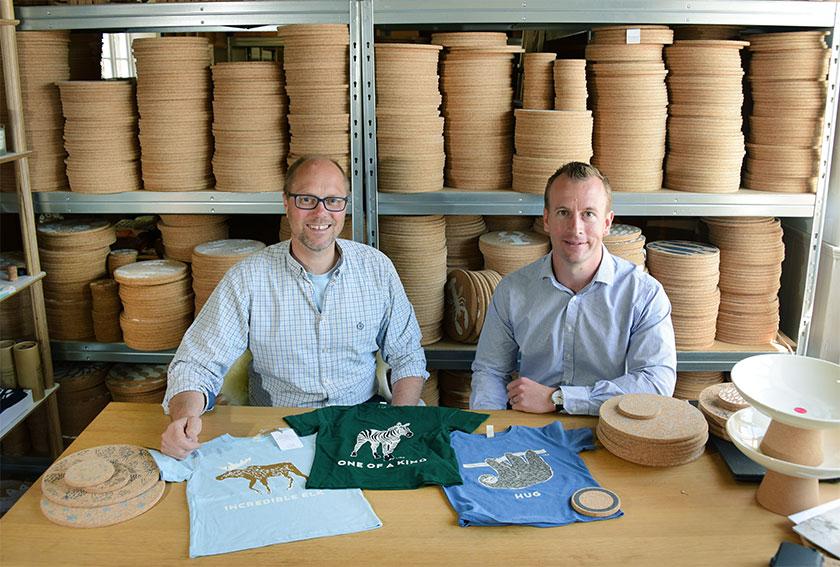 2 men sat at a table showing their cork products and t-shirts with a big stack of cork boards in the background