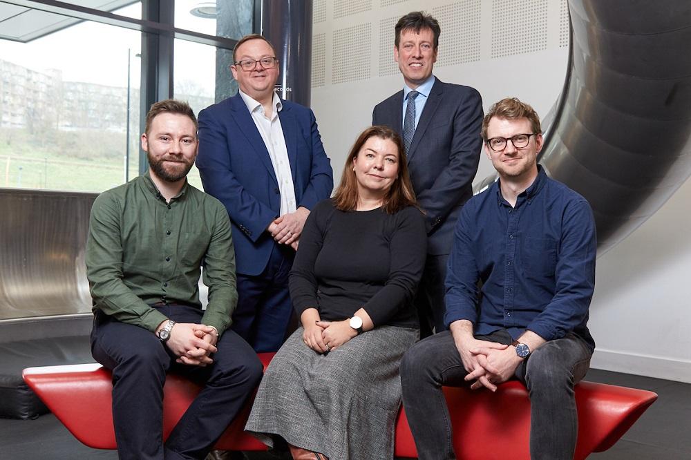 4 men and a woman sat and stood in a meeting room with wall art in the background