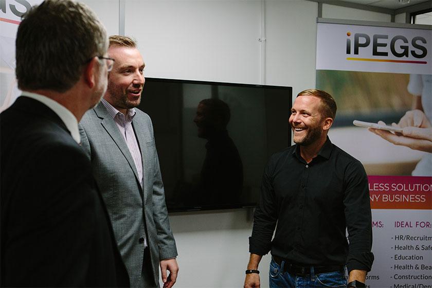 3 men having a conversation in a meeting room with a TV screen on the wall and and iPEGS pop up banner in the background