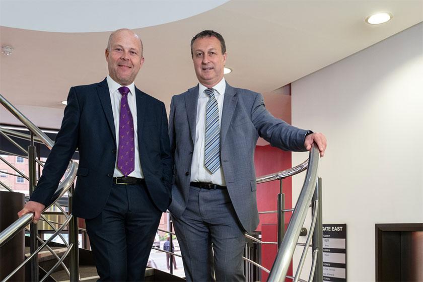 2 men in suits smiling and stood on some stairs inside an office building