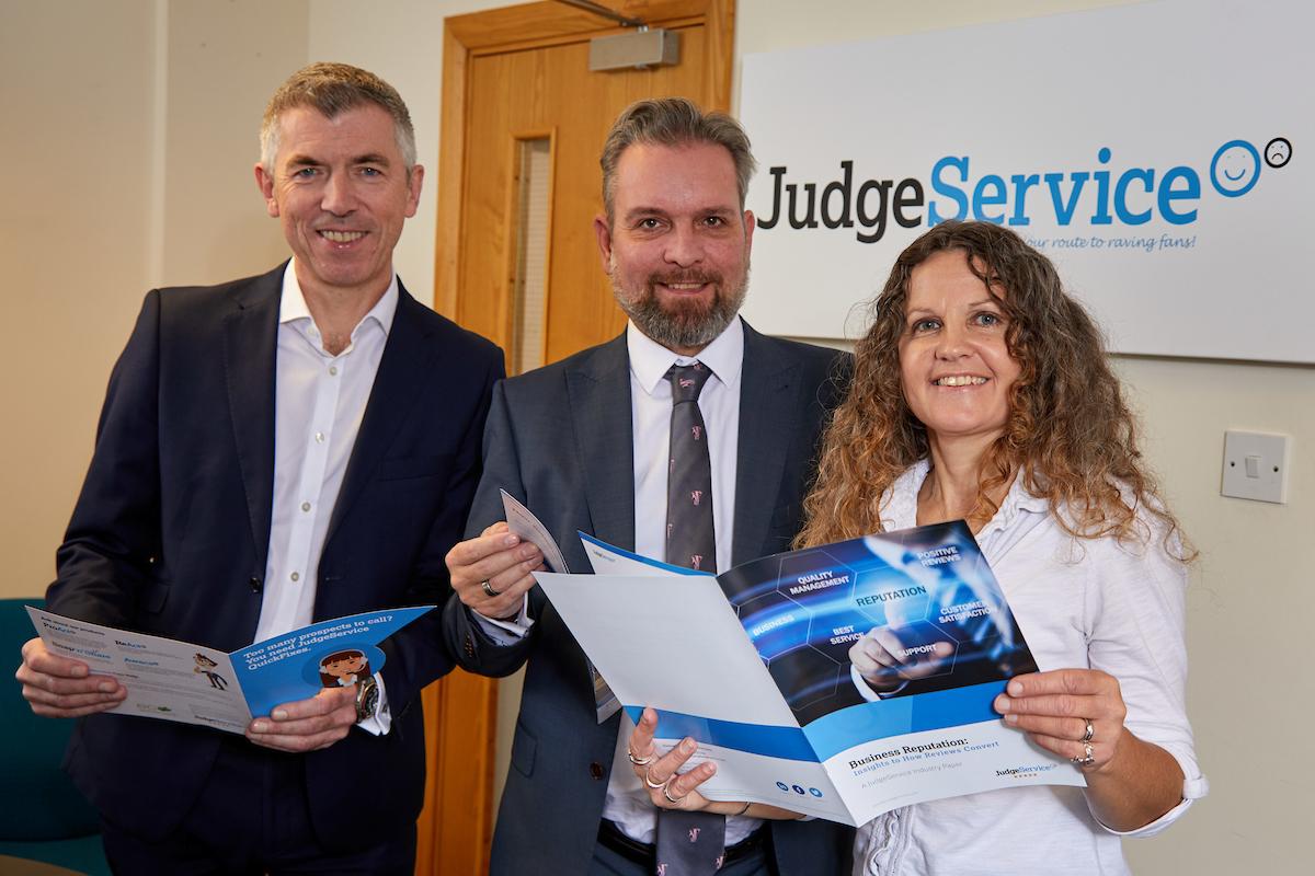 2 men and a woman stood in an office holding some brochures with a Judge Service Research logo on the wall behind them
