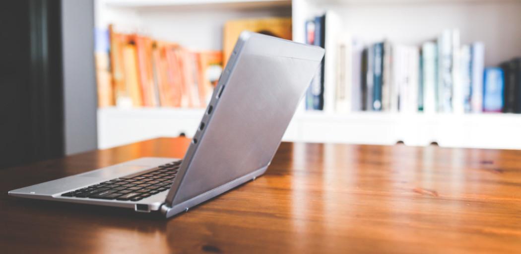 A laptop on a wooden table with a shelf of books in the background