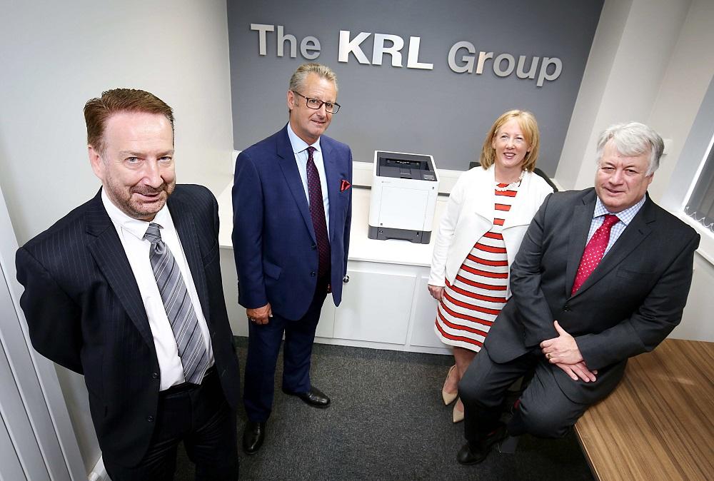 3 men and a woman stood in a meeting room with The KRL Group name on the wall and a printer in between them