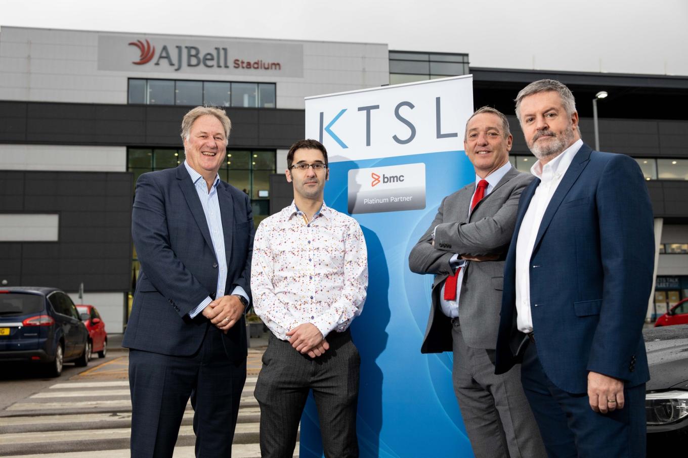 4 men in suits stood in a car park outside of the AJ Bell Stadium with a KTSL pop up banner between them