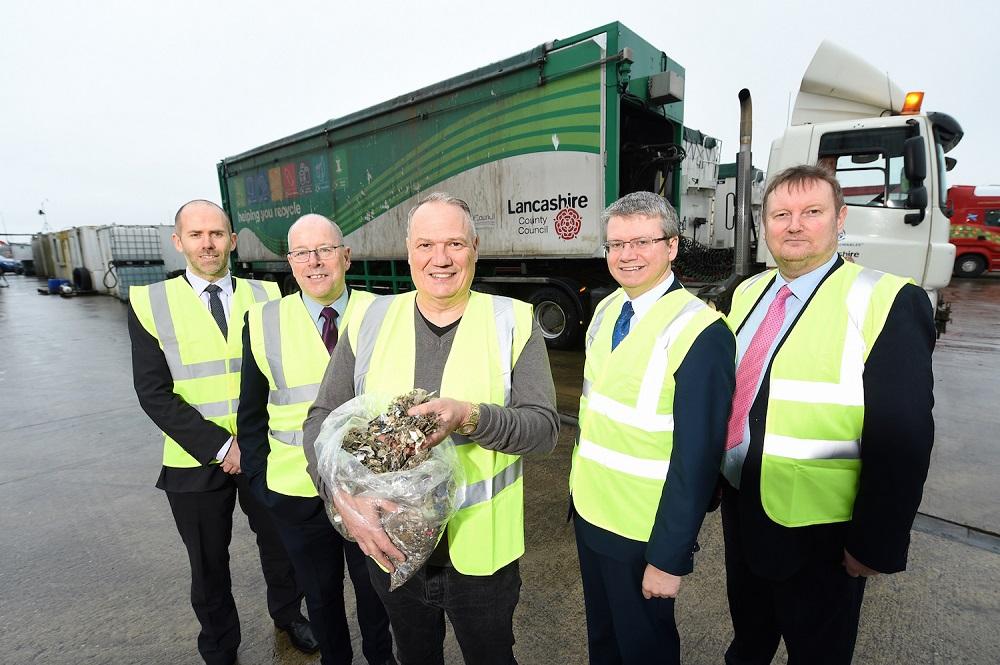 5 men in Hi - Vis jackets stood in a Waste Recycling plot with one man holding some waste in a bag