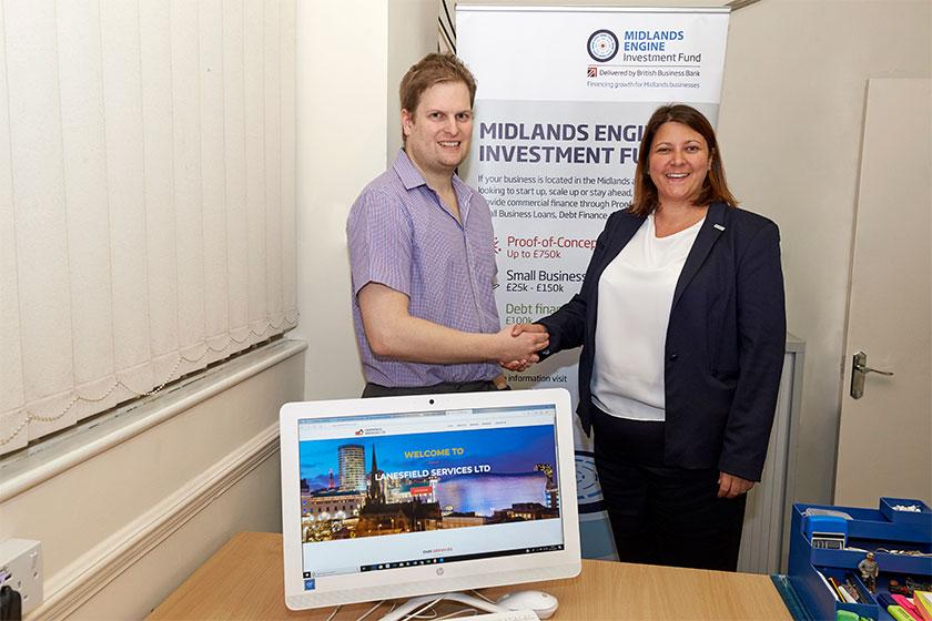 A man and a women shaking hands in a small office with a computer screen on the desk in front