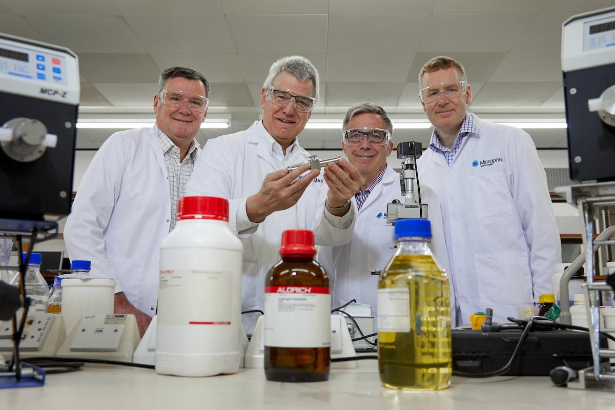 4 men in lab coats and safety goggles in a science lab surrounded by machines and chemicals