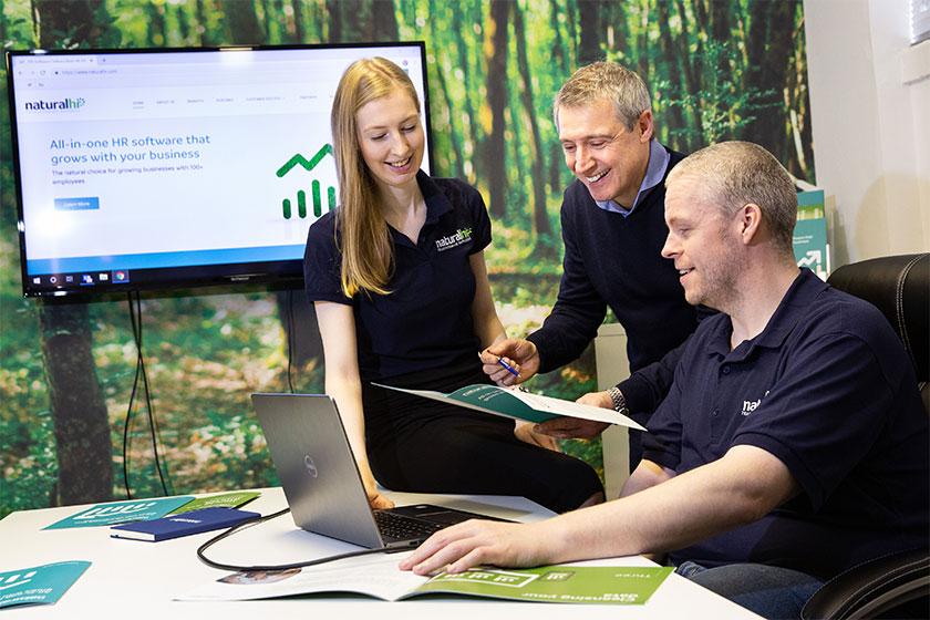 2 men and a woman in a meeting room with a tree themed feature wall looking at a brochure and a laptop