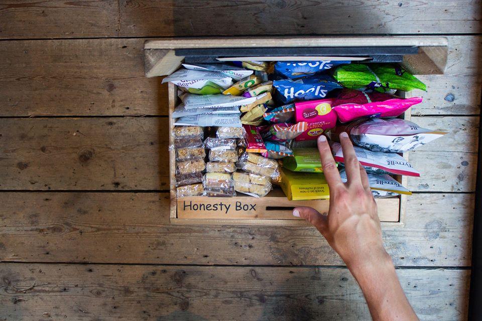 A hand reaching into a wooden box filled with snacks and a slot to donate money