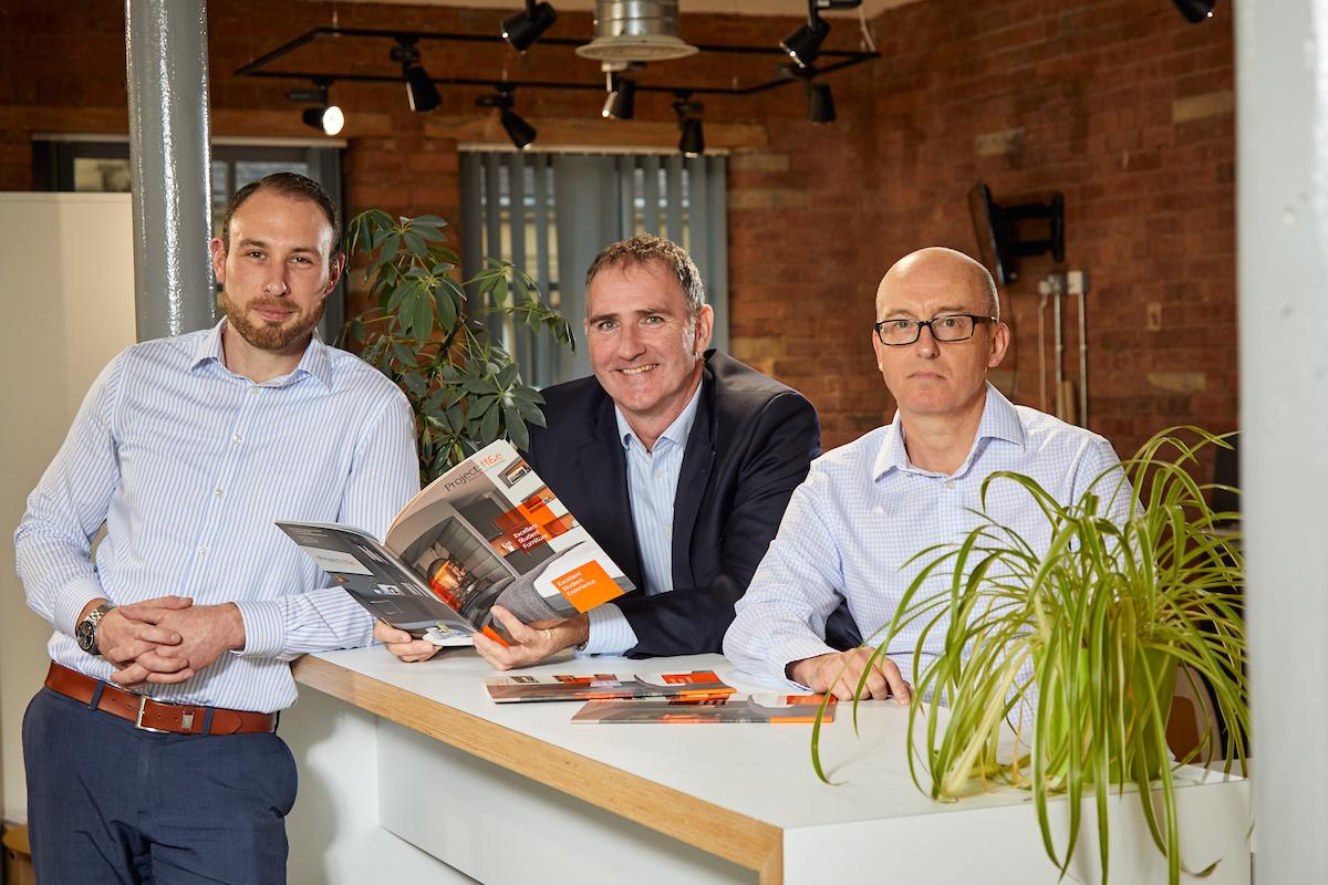 3 men stood around a reception deck with one man holding a Project:ffe brochure