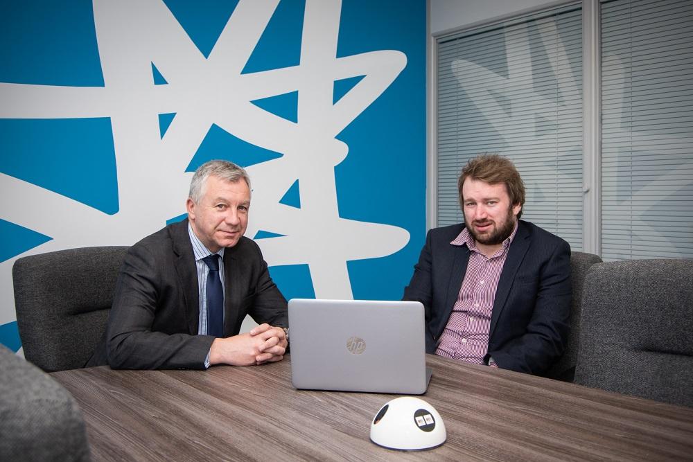 2 men in suits sat at a meeting room table with a laptop in front