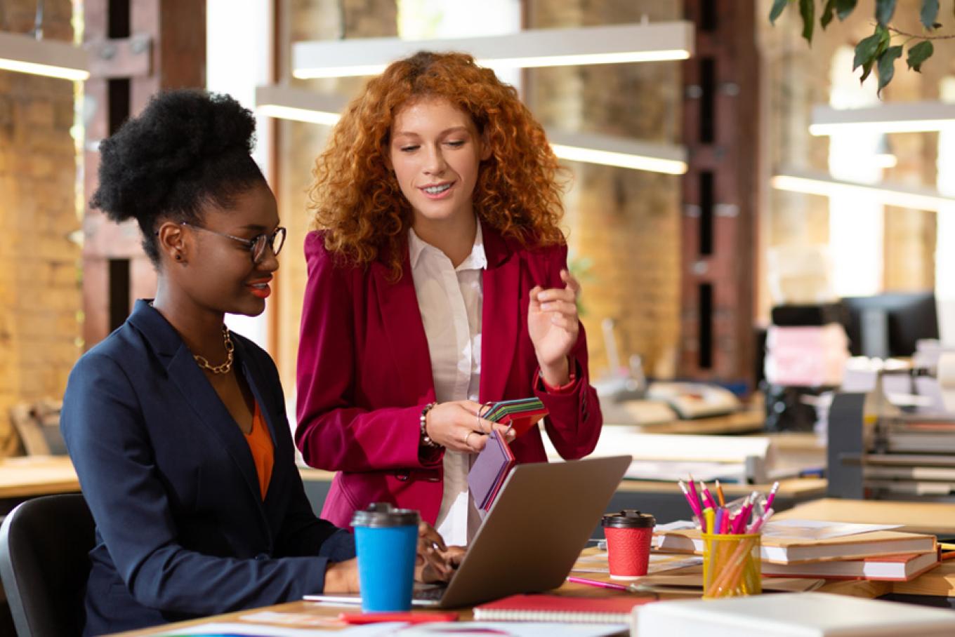 Two businesswomen in an office smiling and talking whilst looking a laptop on a table.