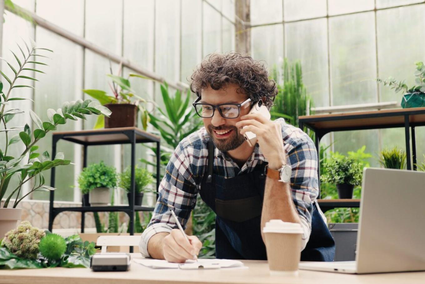 bearded man with glasses on the phone smiling, whilst leaning on a desk with a laptop and coffee on it, in an office with many green plants
